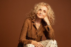 Musician Patty Griffin sits in front of a brown studio background, looking at the camera. She is wearing a brown fringed jacket and studded pants.